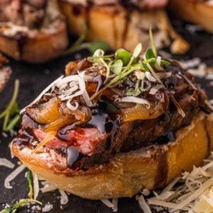 Close up of one steak crostini on slate, the seared crust and pink center of the NY strip visible above the Boursin.