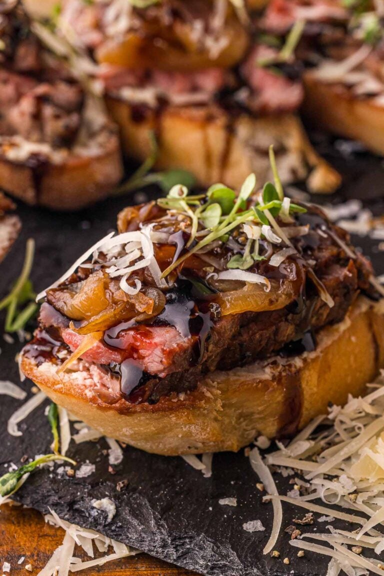 Close up of one steak crostini on slate, the seared crust and pink center of the NY strip visible above the Boursin.