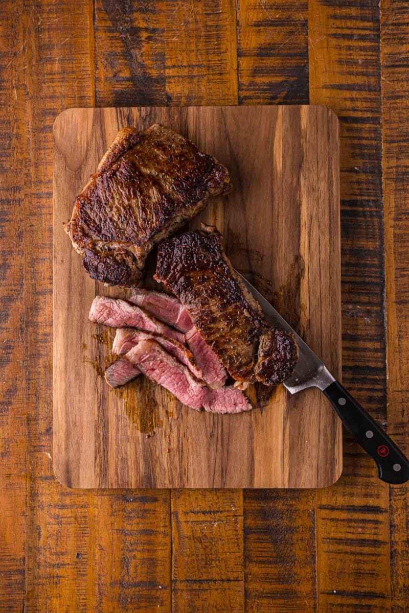 The rested NY strip steaks on a wood cutting board, one sliced thin against the grain to show a medium rare center.