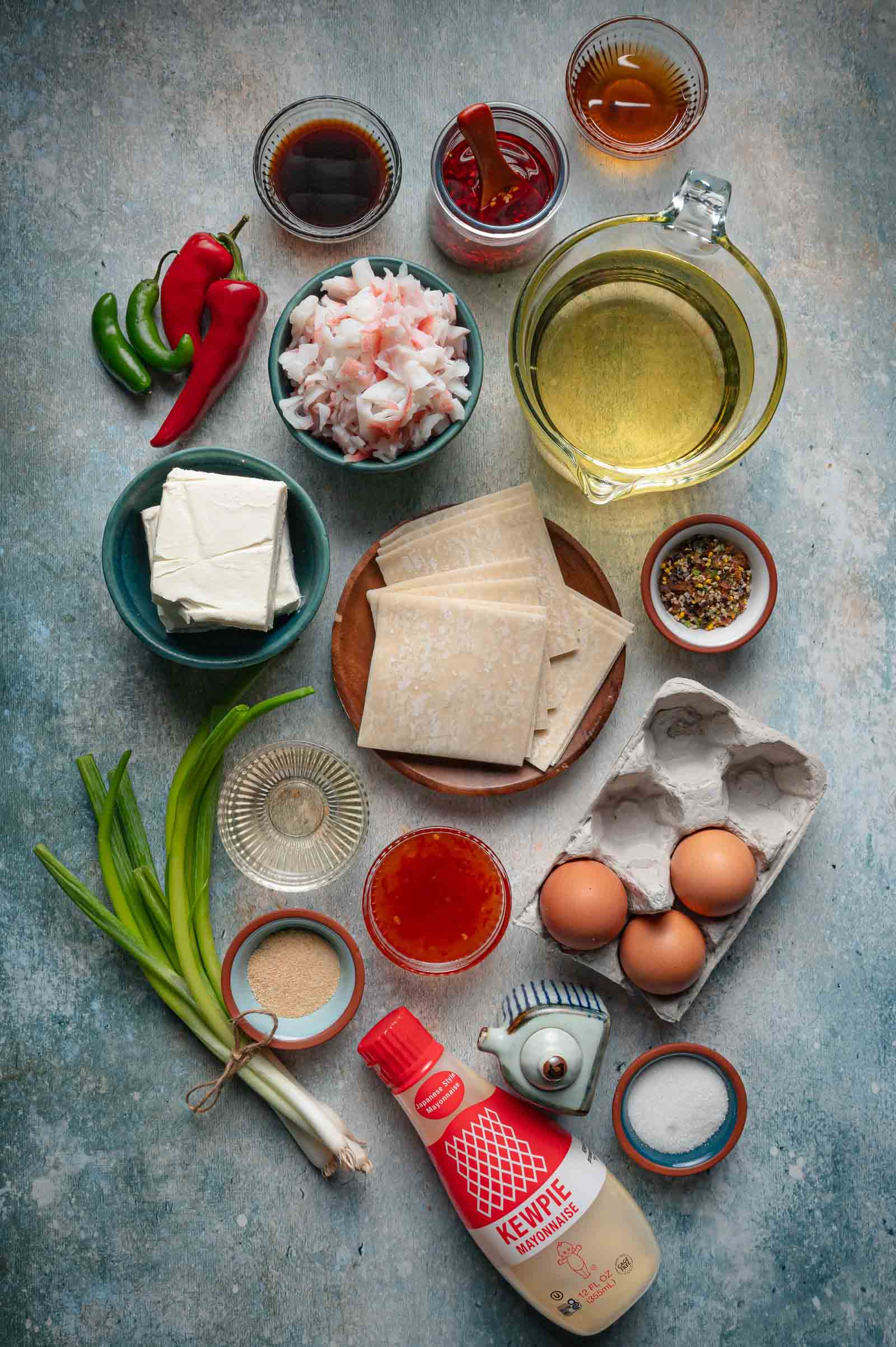 Crab rangoon nachos ingredients laid out: imitation crab, cream cheese, wonton wrappers, scallions, eggs, red and green chilies, sweet chili sauce, soy sauce, chili crisp, frying oil and Kewpie mayonnaise.