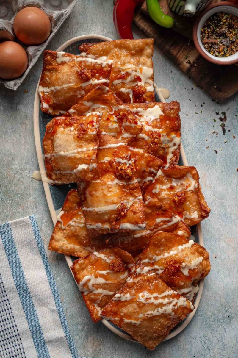 Fried crab rangoon crisps piled on an oval platter with sweet chili sauce, mayo drizzle and chili crisp, before the fresh toppings go on.