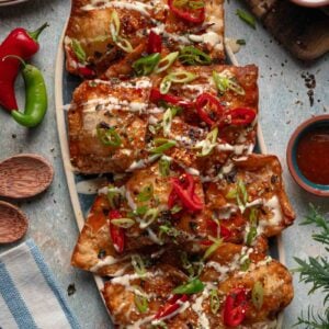 An oval platter of crab rangoon nachos topped with mayo drizzle, chili crisp, sliced red and green chilies and scallions.