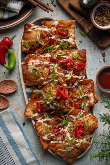 An oval platter of crab rangoon nachos topped with mayo drizzle, chili crisp, sliced red and green chilies and scallions.