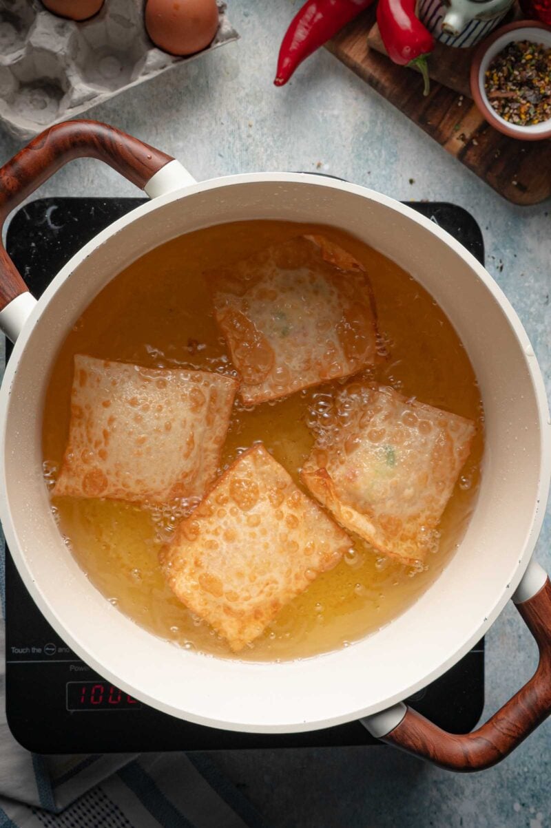 Four sealed wonton packets frying in hot oil, bubbling and turning golden at the edges.