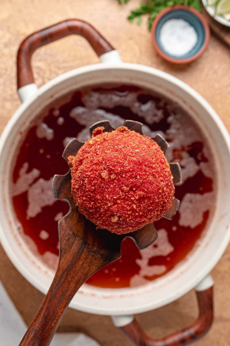 A fried birria ball lifted on a wooden slotted spoon from a pot of frying oil stained deep red by the Hot Cheeto crust.