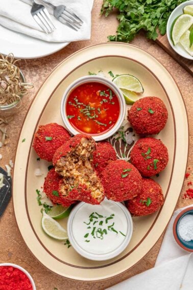 A platter of Hot Cheeto birria balls with one split open to show the beef, rice and melted Oaxaca cheese inside, consomรฉ and crema alongside.