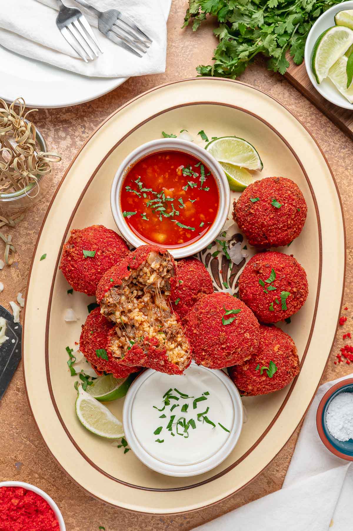 A platter of Hot Cheeto birria balls with one split open to show the beef, rice and melted Oaxaca cheese inside, consomé and crema alongside.