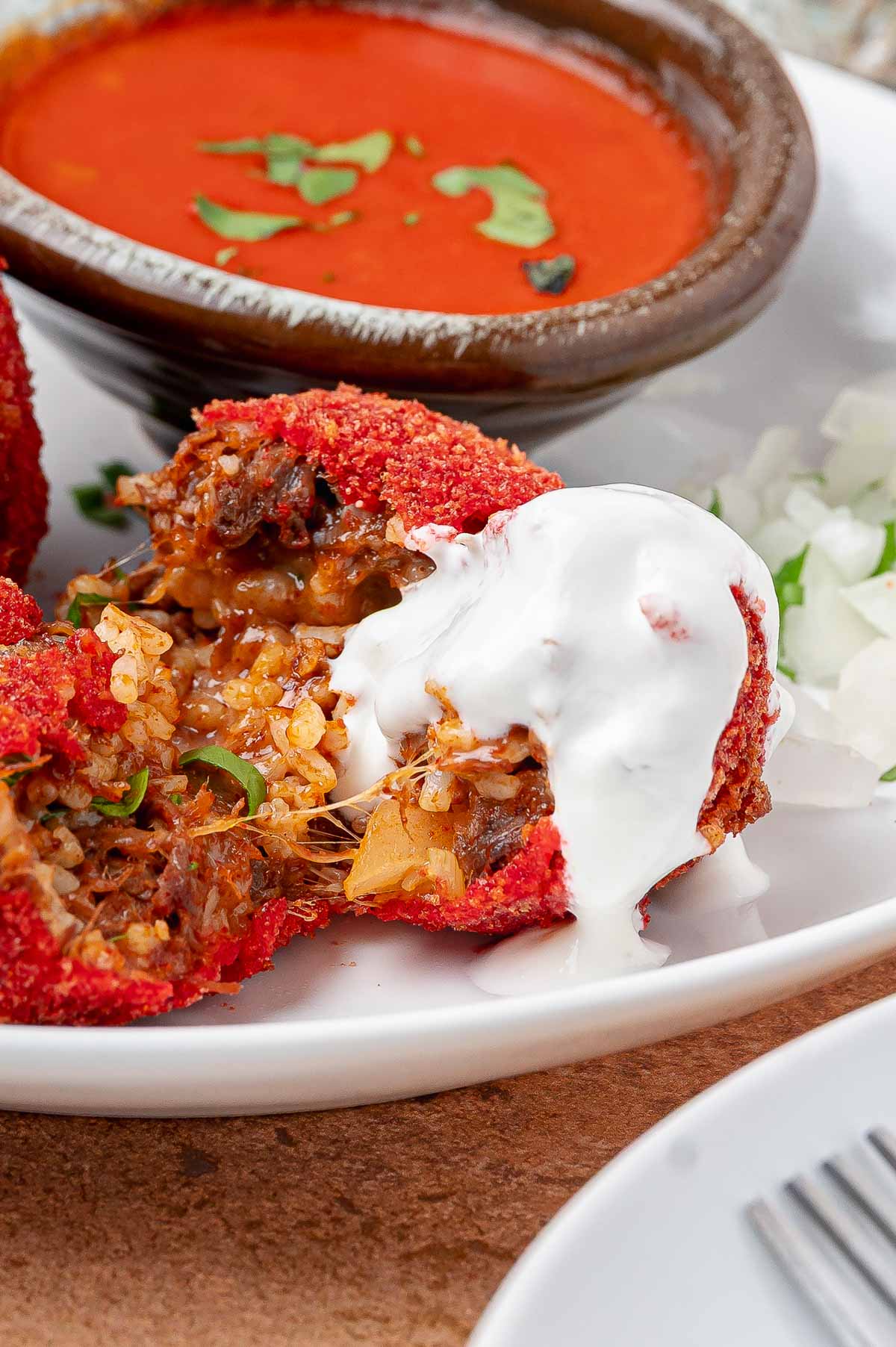 A split Hot Cheeto birria ball on a plate under a pour of Mexican crema, with diced white onion and a clay bowl of birria consomé.