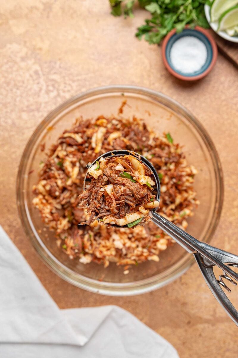 A spring loaded scoop lifting a portion of the birria and rice filling out of the mixing bowl to portion the balls evenly.