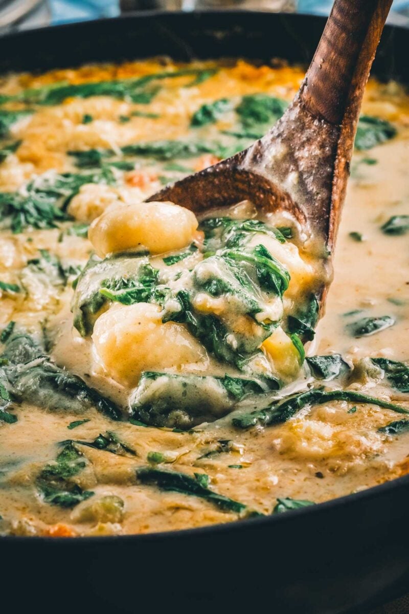 A wooden spoon lifting potato gnocchi and wilted spinach out of the creamy broth of the finished chicken gnocchi soup.
