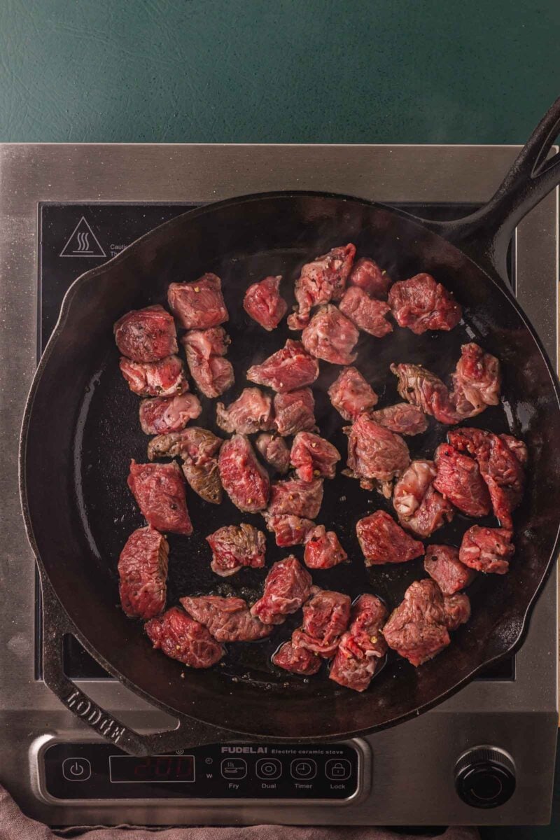 Raw sirloin steak bites seasoned with salt and cracked pepper, spread in a single layer in a hot cast iron skillet to sear.