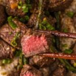 Macro of a miso butter steak bite held in chopsticks, cut to show the pink center, over the bowl of rice and green beans.