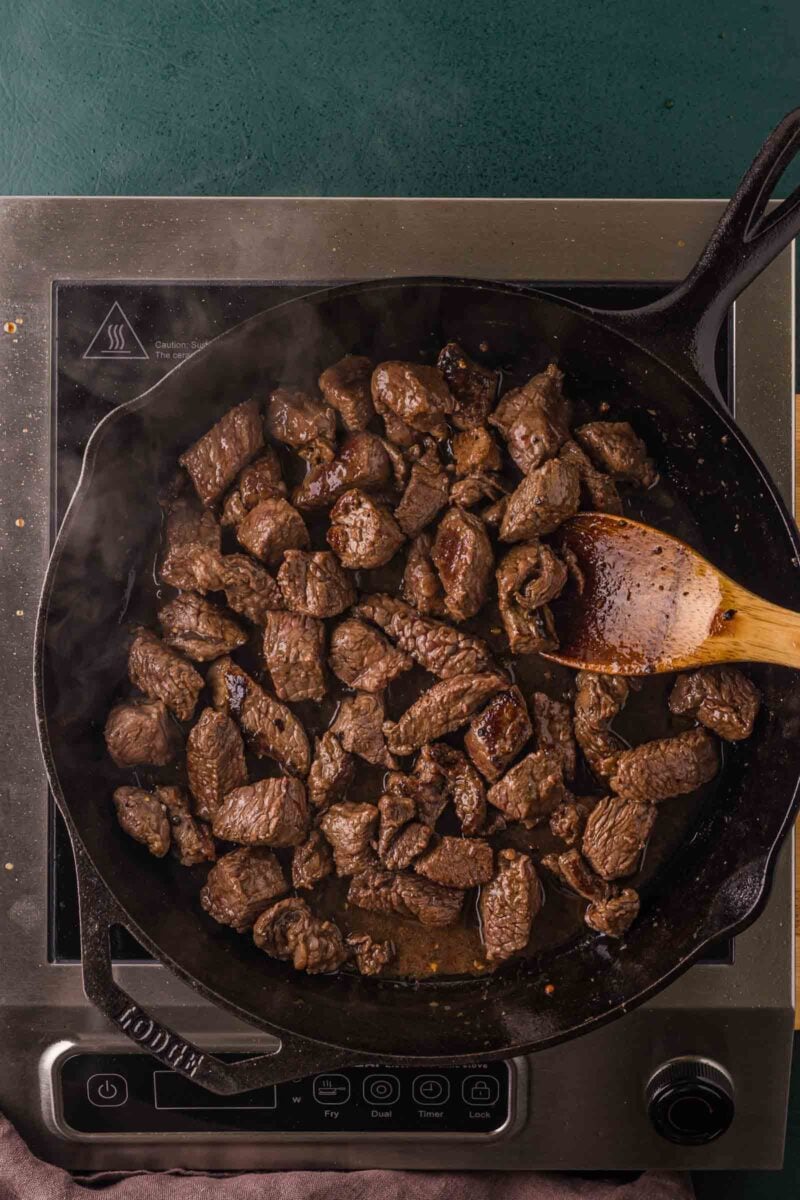 Sirloin steak bites turned in the cast iron skillet with a wooden spoon, crust seared deep brown all over.