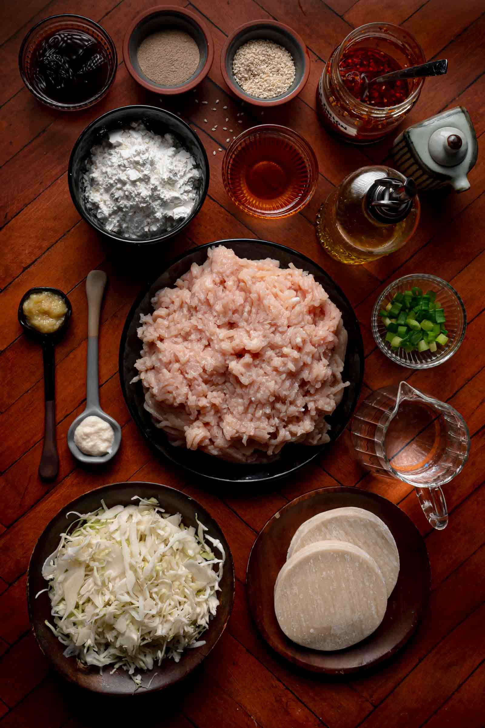 Top-down view of raw dumpling ingredients including ground chicken, cabbage, green onions, sauces, seasonings, dumpling wrappers, and liquid in bowls on a wooden surface.