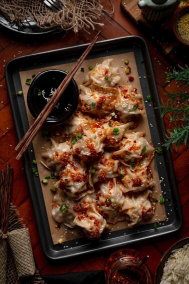 A tray of cooked blanket dumplings topped with chili oil, chopped green onions, and sesame seeds, served with a small bowl of dipping sauce and chopsticks on the side.