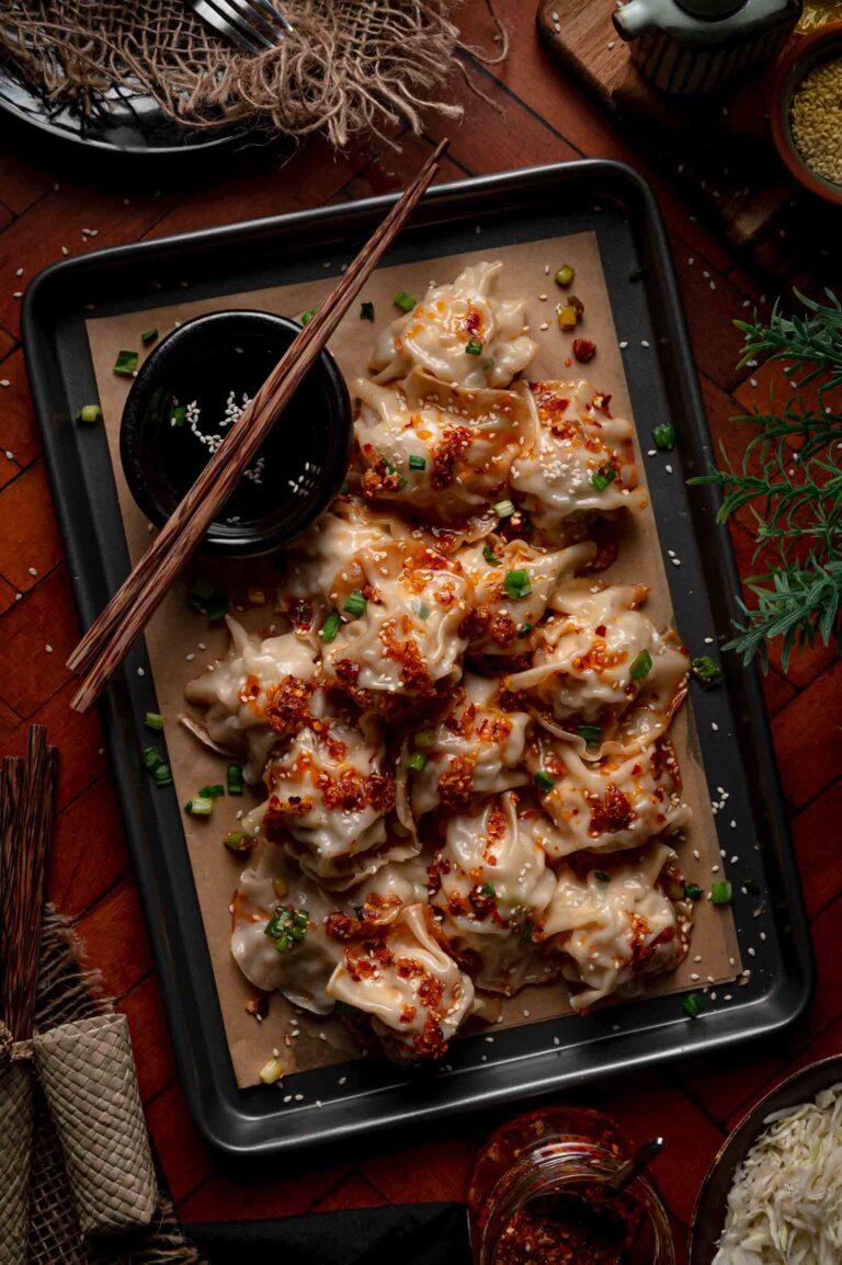 A tray of cooked blanket dumplings topped with chili oil, chopped green onions, and sesame seeds, served with a small bowl of dipping sauce and chopsticks on the side.