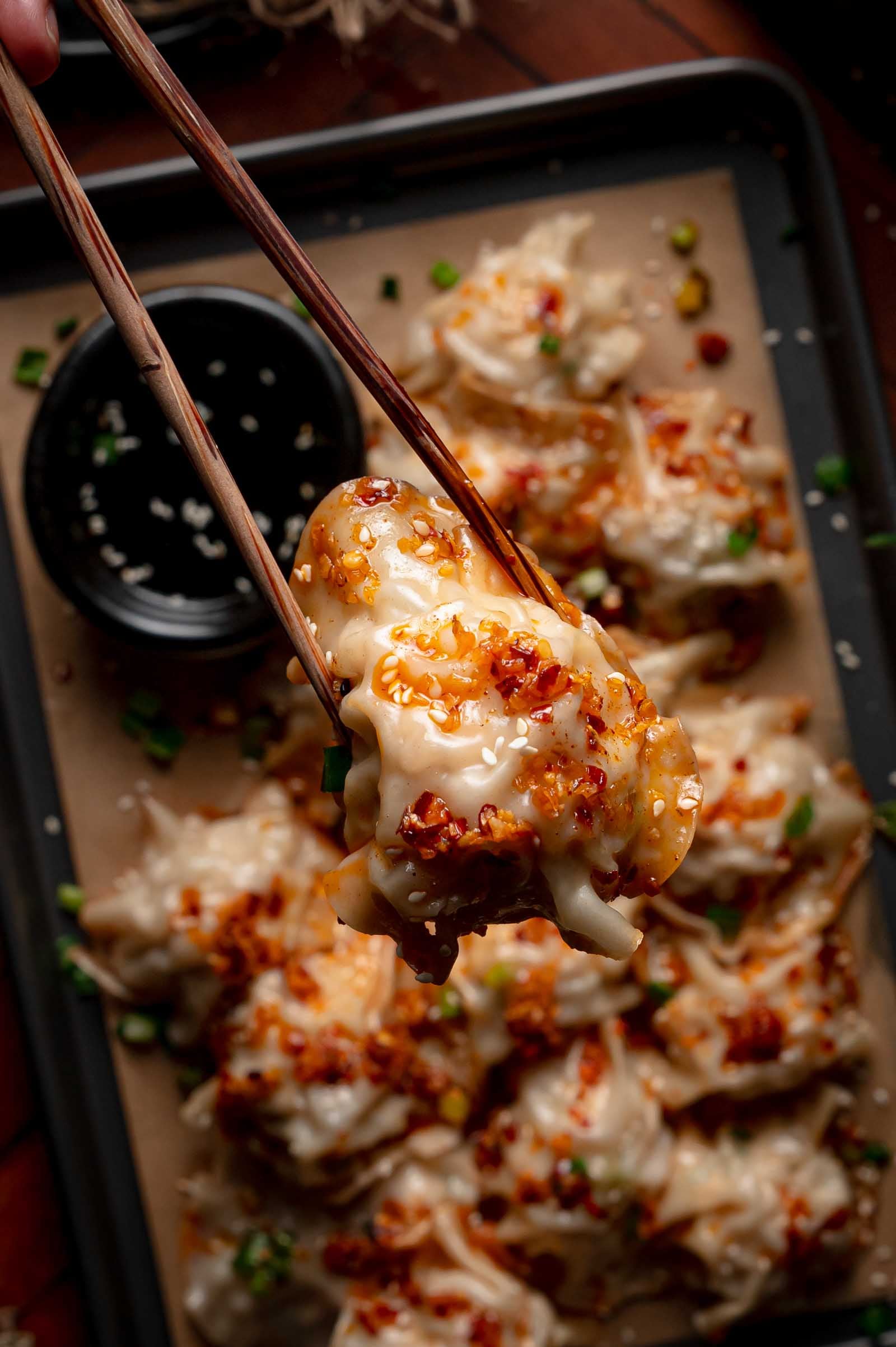 Chopsticks hold a cooked blanket dumpling topped with chili oil above a tray of more dumplings, with a small bowl of dipping sauce nearby.