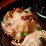 Close-up of dumplings topped with chili flakes, sesame seeds, and chopped green onions on a black plate.