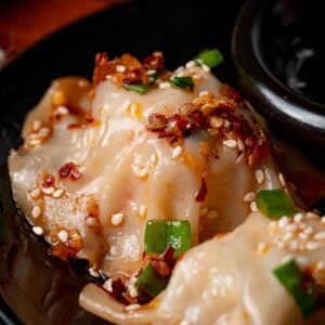 Close-up of dumplings topped with chili flakes, sesame seeds, and chopped green onions on a black plate.