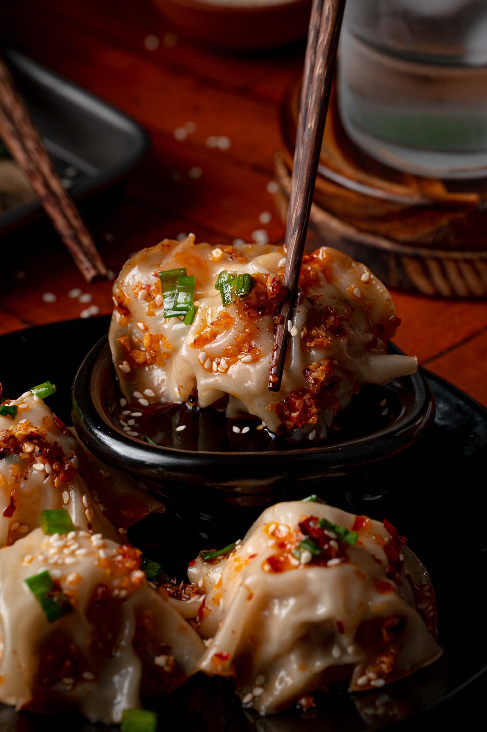 Three blanket dumplings topped with chili oil, sesame seeds, and chopped green onions, with chopsticks holding one dumpling over a small dipping bowl.