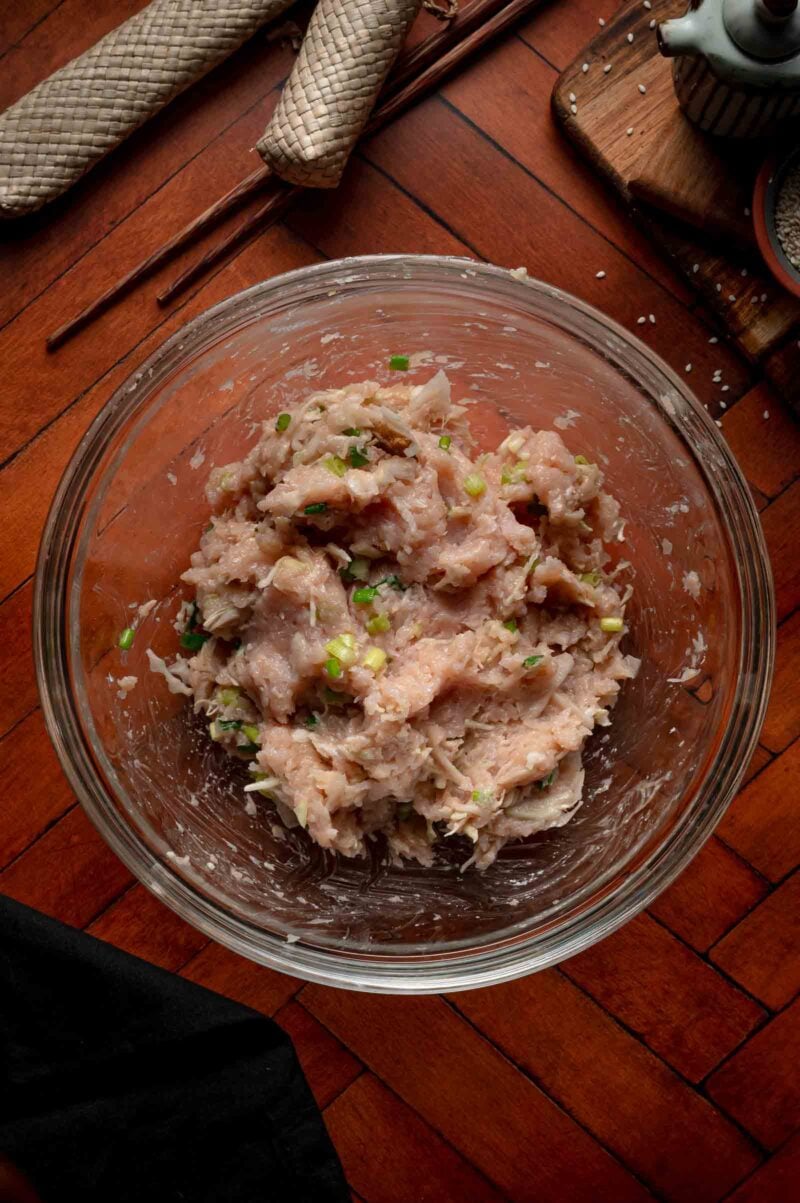 A glass bowl filled with raw ground chicken mixed with chopped green onions sits on a wooden table.