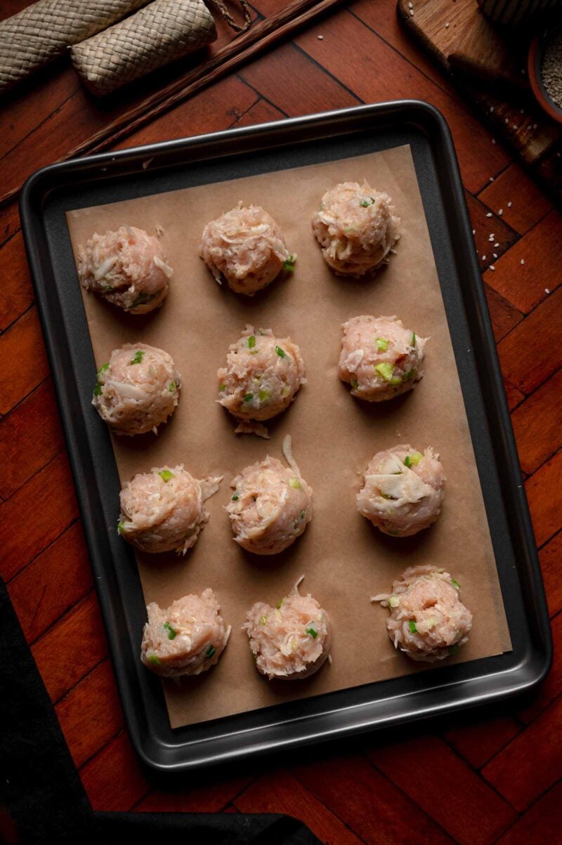 Twelve raw chicken dumpling filling portions arranged on a parchment-lined baking tray, ready for cooking, on a wooden surface.