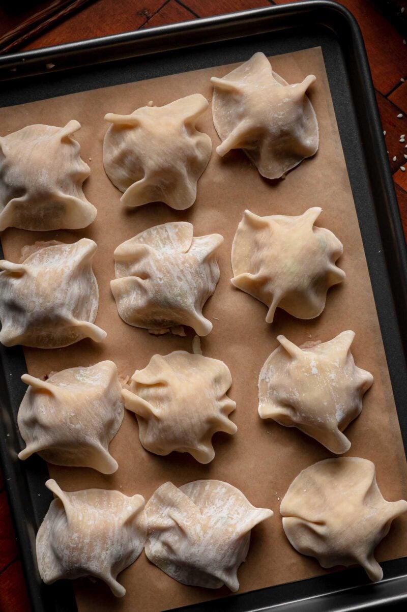 Twelve uncooked blanket dumplings with edges draped over the filling are arranged on a baking sheet lined with parchment paper.