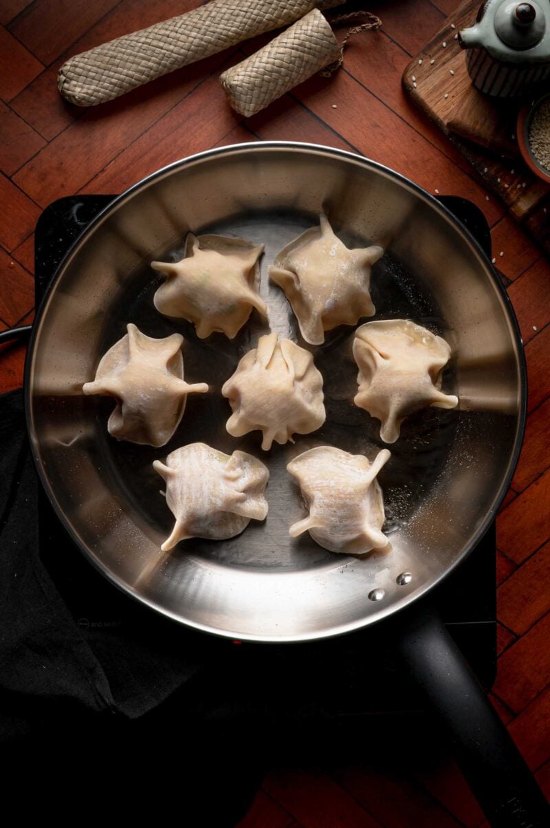 Seven uncooked blanket dumplings with pleated tops are arranged in a nonstick frying pan on a stovetop, ready to be cooked. Various kitchen items are visible in the background.
