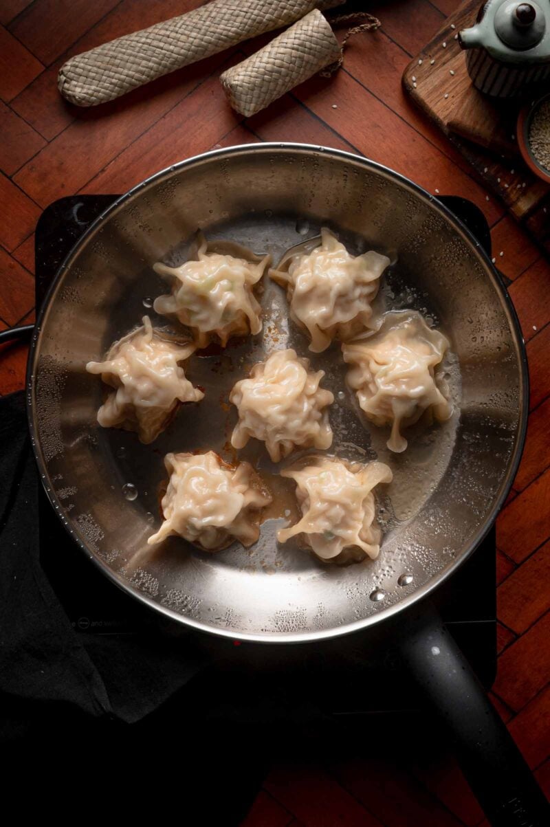 Seven blanket dumplings are being cooked in a metal pan on a stovetop, with various kitchen items and a wooden surface visible in the background.