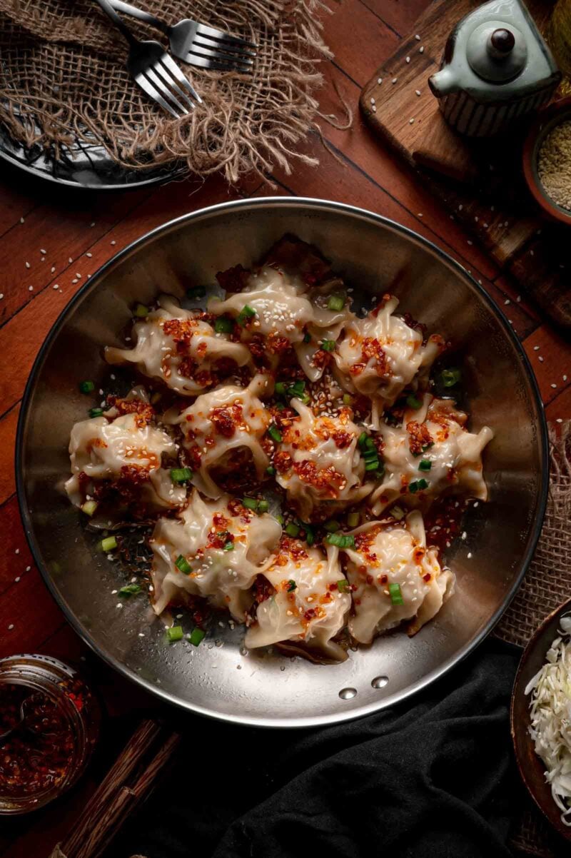 A bowl of steamed blanket dumplings topped with chili oil, chopped green onions, and sesame seeds sits on a wooden table with utensils and condiments nearby.