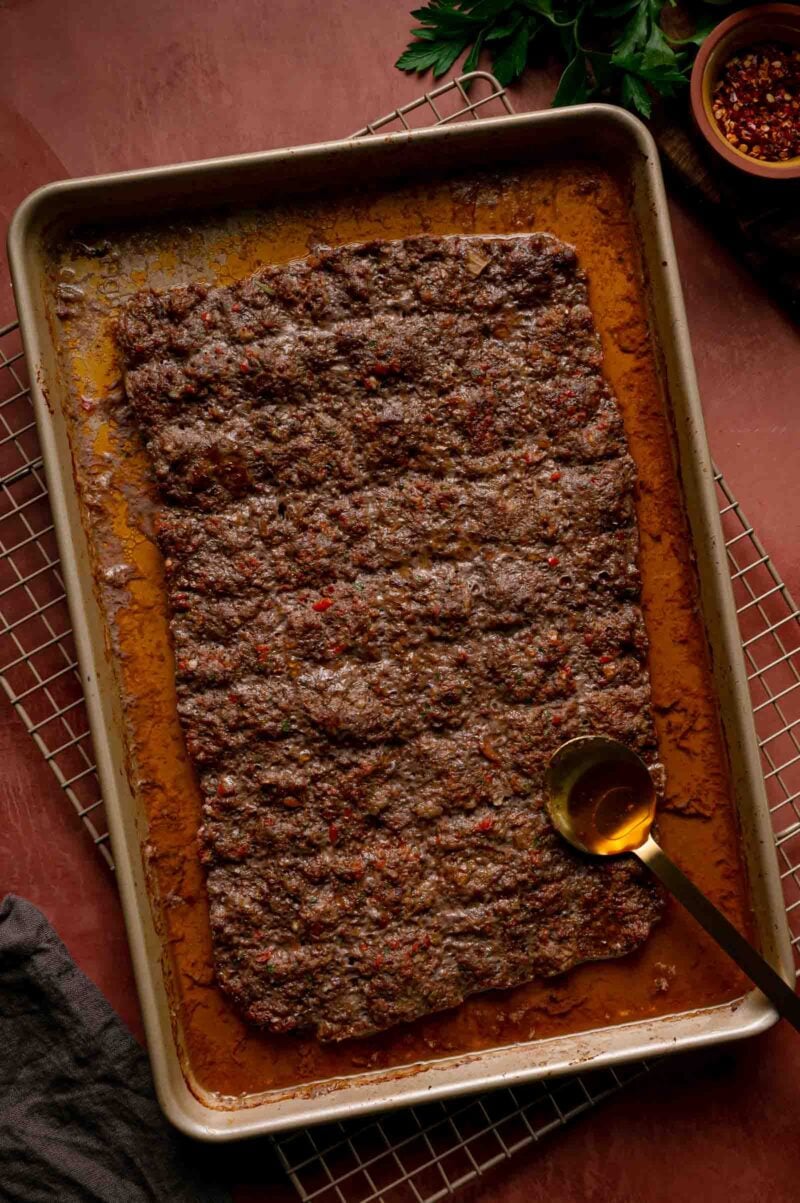 The baked kebab slab in the sheet pan with rendered fat pooled at the edges and a spoon for draining it off.