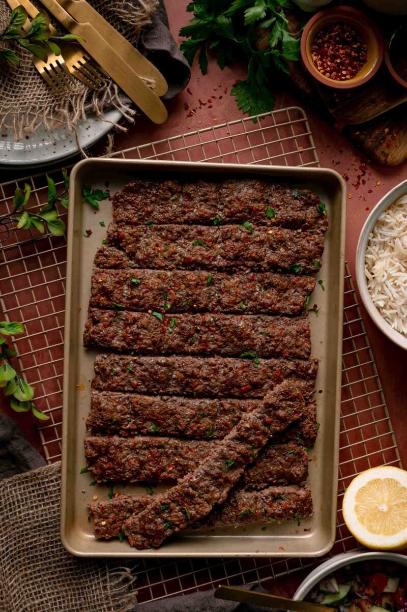 Baked sheet pan kebabs cut into strips and scattered with parsley, resting on a rack beside rice and lemon.