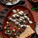 Sheet pan kebab strips on a wooden platter with herbed yogurt sauce, chopped cucumber and tomato salad, and flatbread.