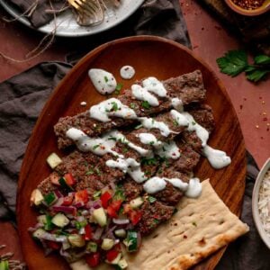 Sheet pan kebab strips on a wooden platter with herbed yogurt sauce, chopped cucumber and tomato salad, and flatbread.