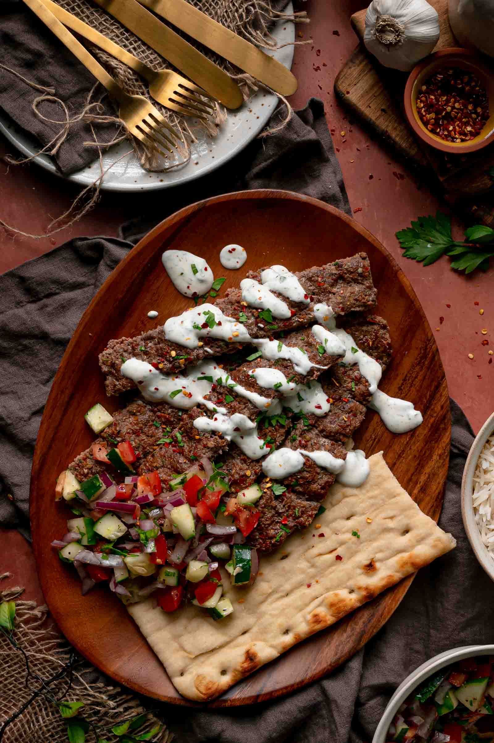 Sheet pan kebab strips on a wooden platter with herbed yogurt sauce, chopped cucumber and tomato salad, and flatbread.