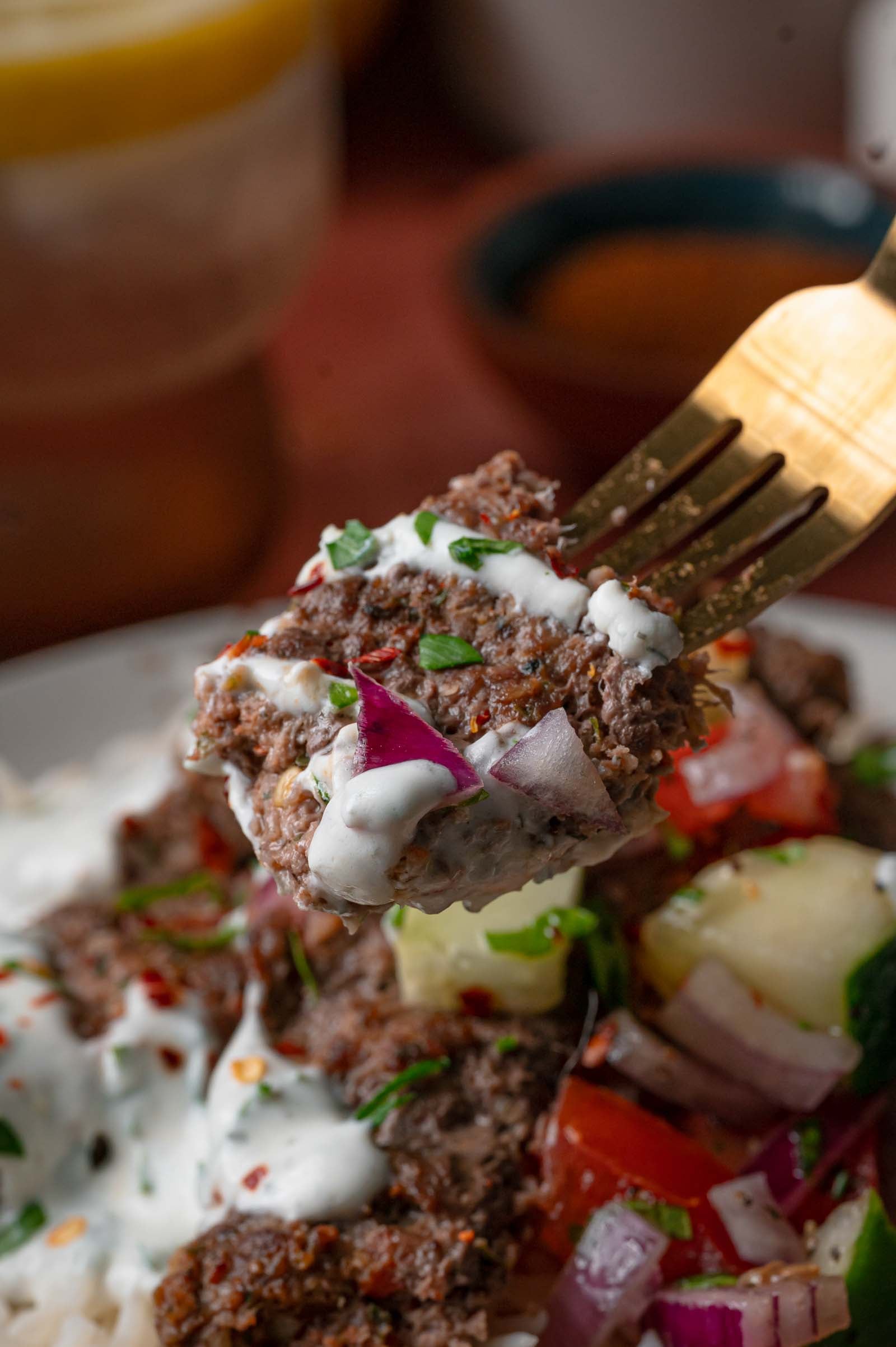 Close-up of a fork holding a sheet pan kebab piece topped with yogurt sauce, red onion and parsley.