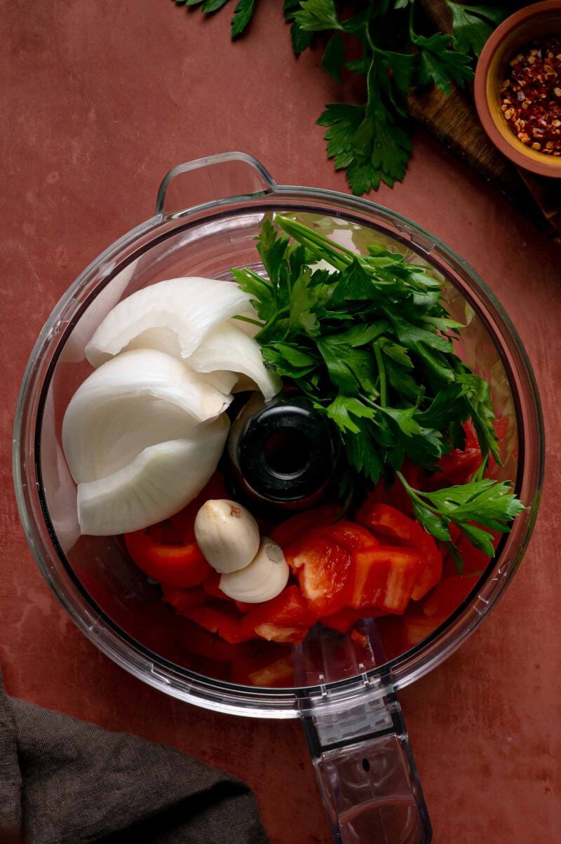 Onion, garlic, red bell pepper and parsley in a food processor bowl before pulsing.