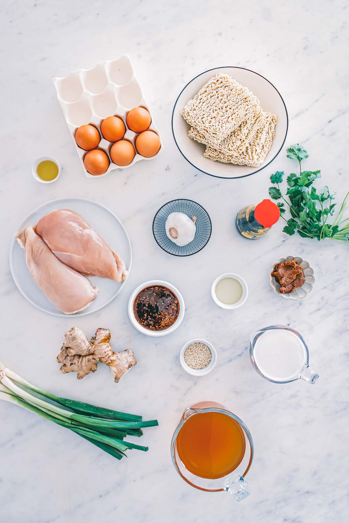 Ingredients for teriyaki chicken ramen: raw chicken breasts, ramen blocks, eggs, red miso, coconut milk, chicken broth, soy sauce, mirin, ginger, garlic, green onion, cilantro and sesame seeds.