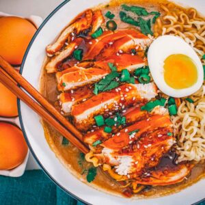 Overhead bowl of teriyaki chicken ramen, sliced chicken glazed in teriyaki beside a halved jammy egg and curled ramen noodles.