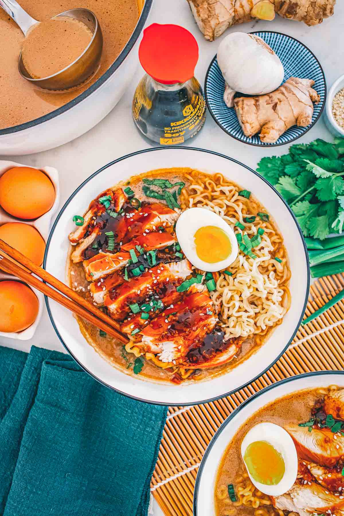 Teriyaki chicken ramen with the pot of broth, soy sauce, ginger, garlic and cilantro around it, chopsticks resting on the bowl.