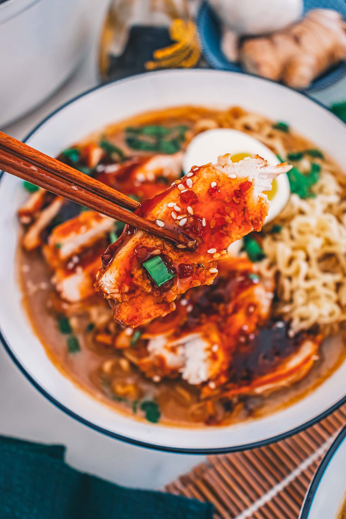 Chopsticks lifting a slice of teriyaki-glazed chicken breast topped with sesame seeds and green onion over the ramen bowl.