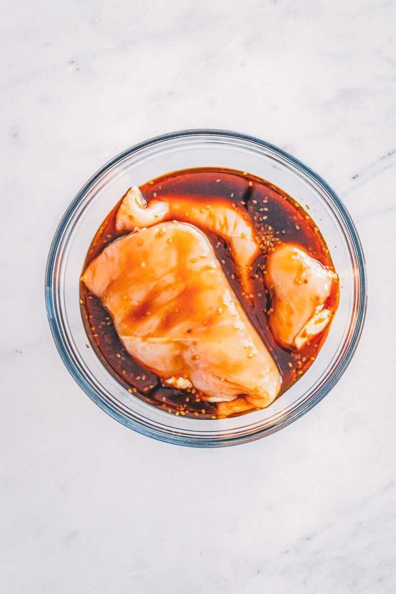 Two boneless skinless chicken breasts marinating in soy sauce and teriyaki in a glass bowl.