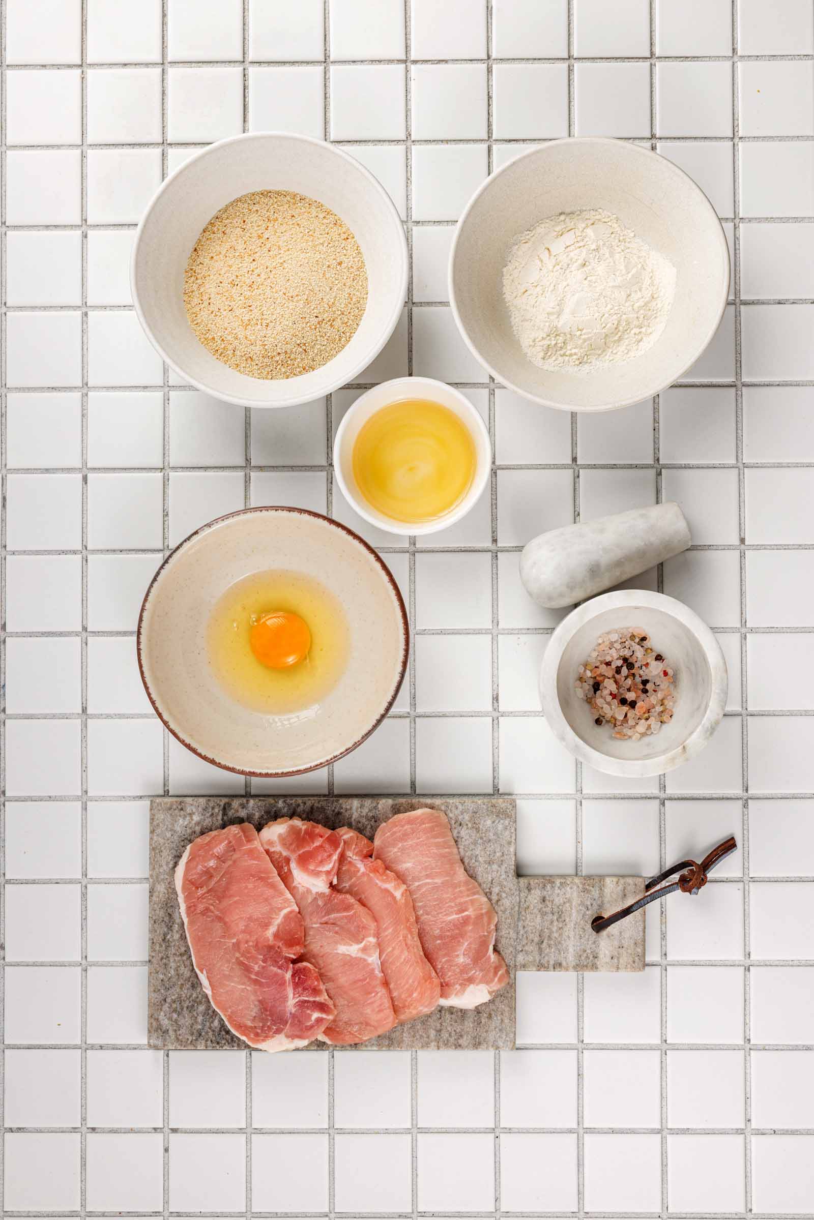 Ingredients for a tonkatsu sandwich: three raw pork loin cutlets on a stone board with bowls of breadcrumbs, flour, egg and cracked peppercorns.