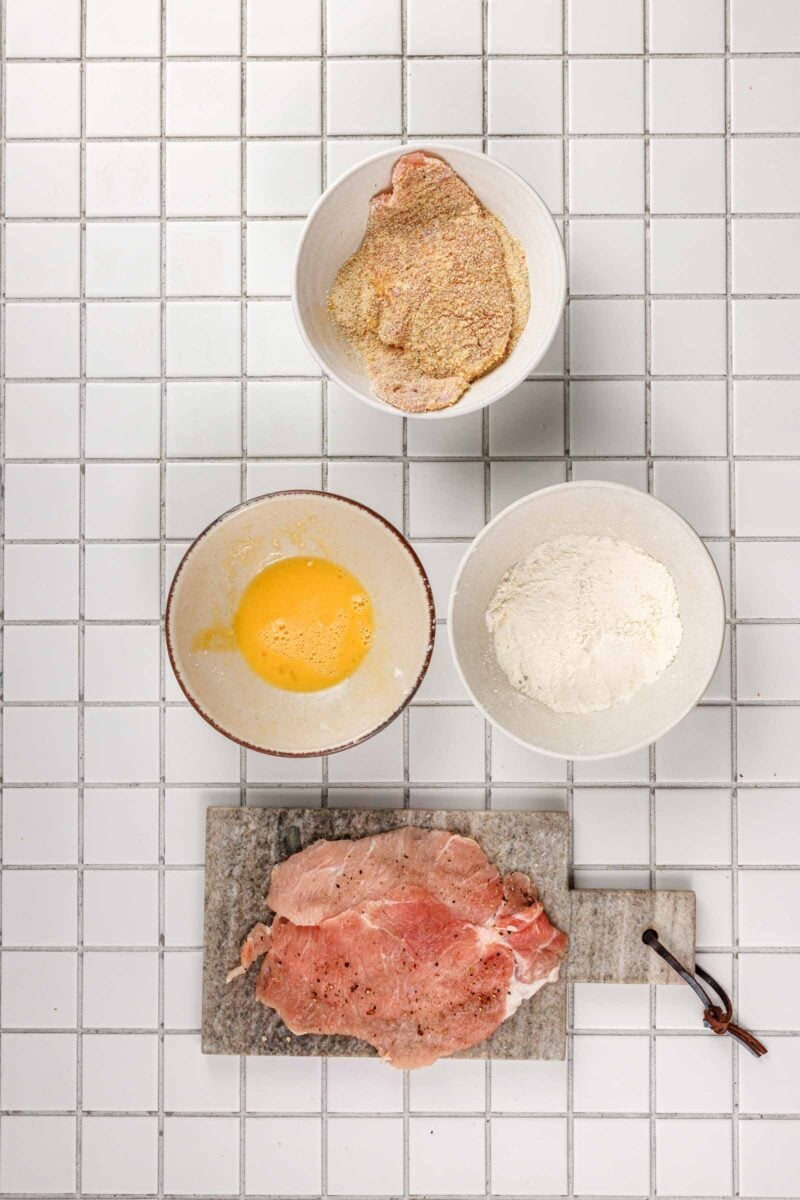 A pork cutlet coated in seasoned breadcrumbs beside bowls of flour and beaten egg, with a second pounded and peppered cutlet waiting on the board.
