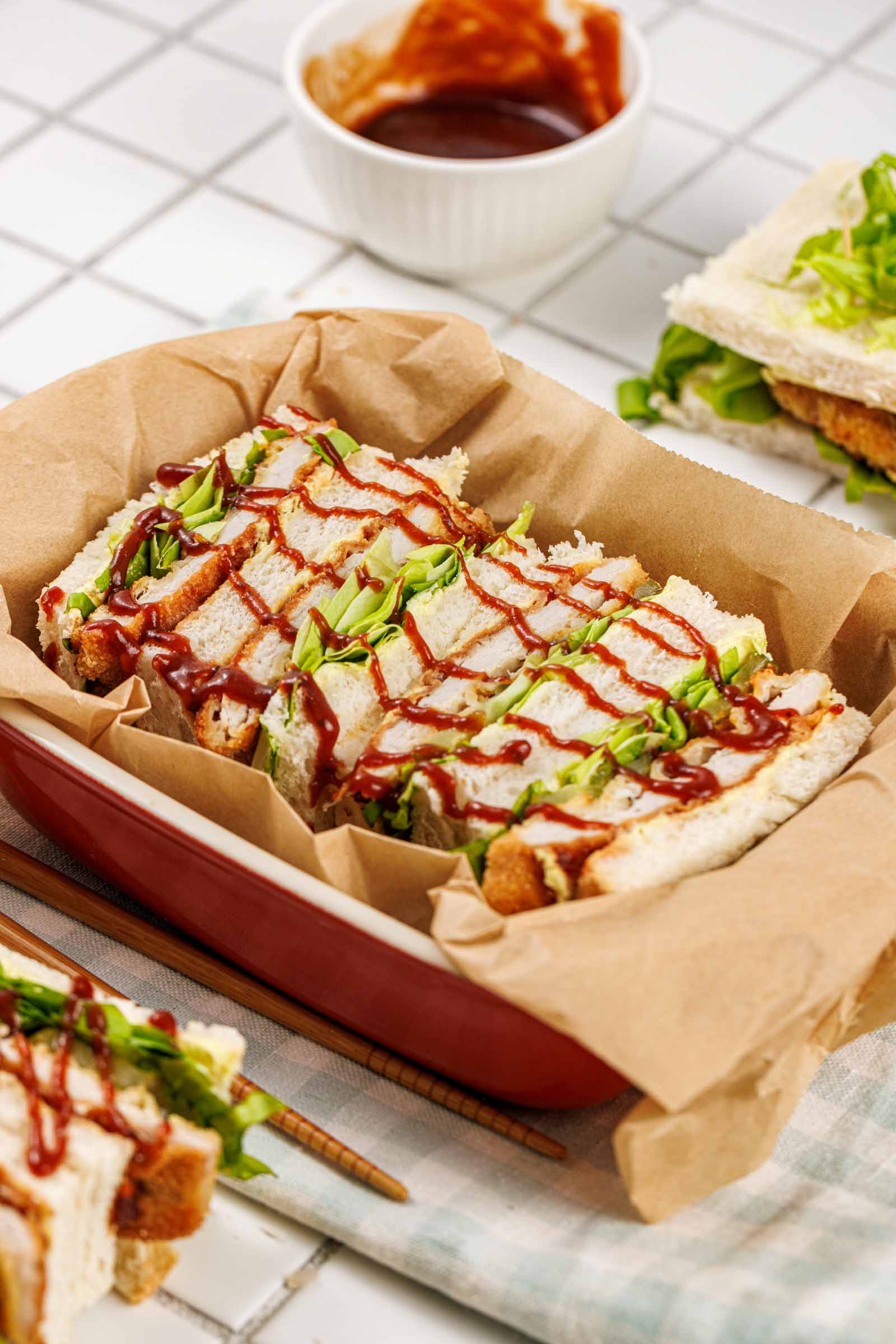 Tonkatsu sandwiches in a red parchment lined dish, tonkatsu sauce drizzled over the top in a lattice, with a bowl of sauce and chopsticks alongside.