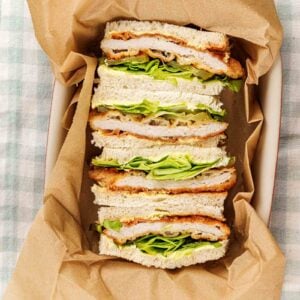 Tonkatsu sandwiches stood on end in a parchment lined dish, breaded pork cutlet, chopped greens and pickle slices between crustless white bread.