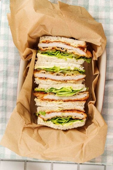 Tonkatsu sandwiches stood on end in a parchment lined dish, breaded pork cutlet, chopped greens and pickle slices between crustless white bread.