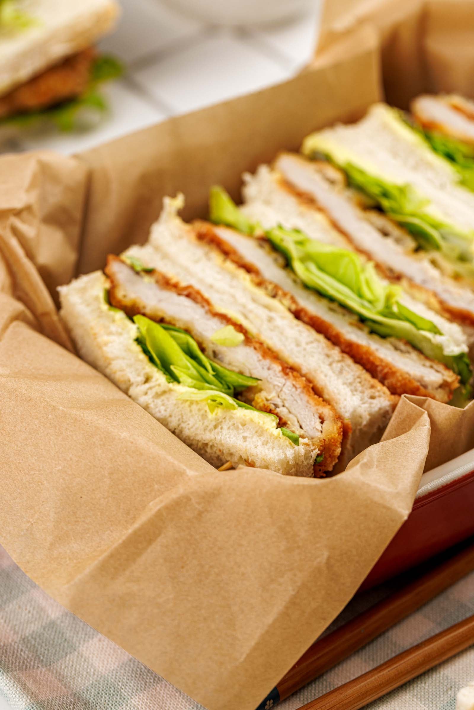 Tonkatsu sandwich halves in a parchment lined dish, the breaded pork cutlet, chopped greens and pickle slices visible against the soft white bread.