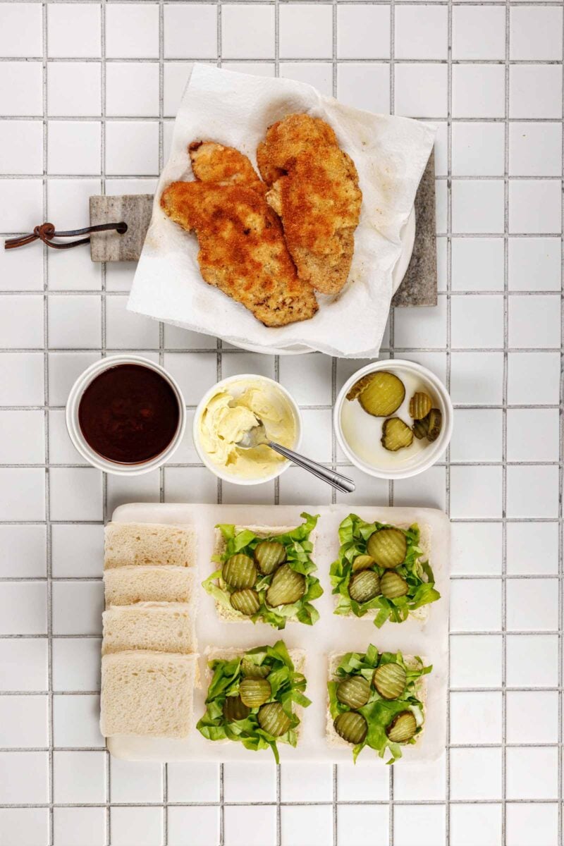 Fried pork cutlets draining on paper towel beside bowls of tonkatsu sauce, Kewpie mayo and pickle chips, with crustless bread laid out with chopped greens.