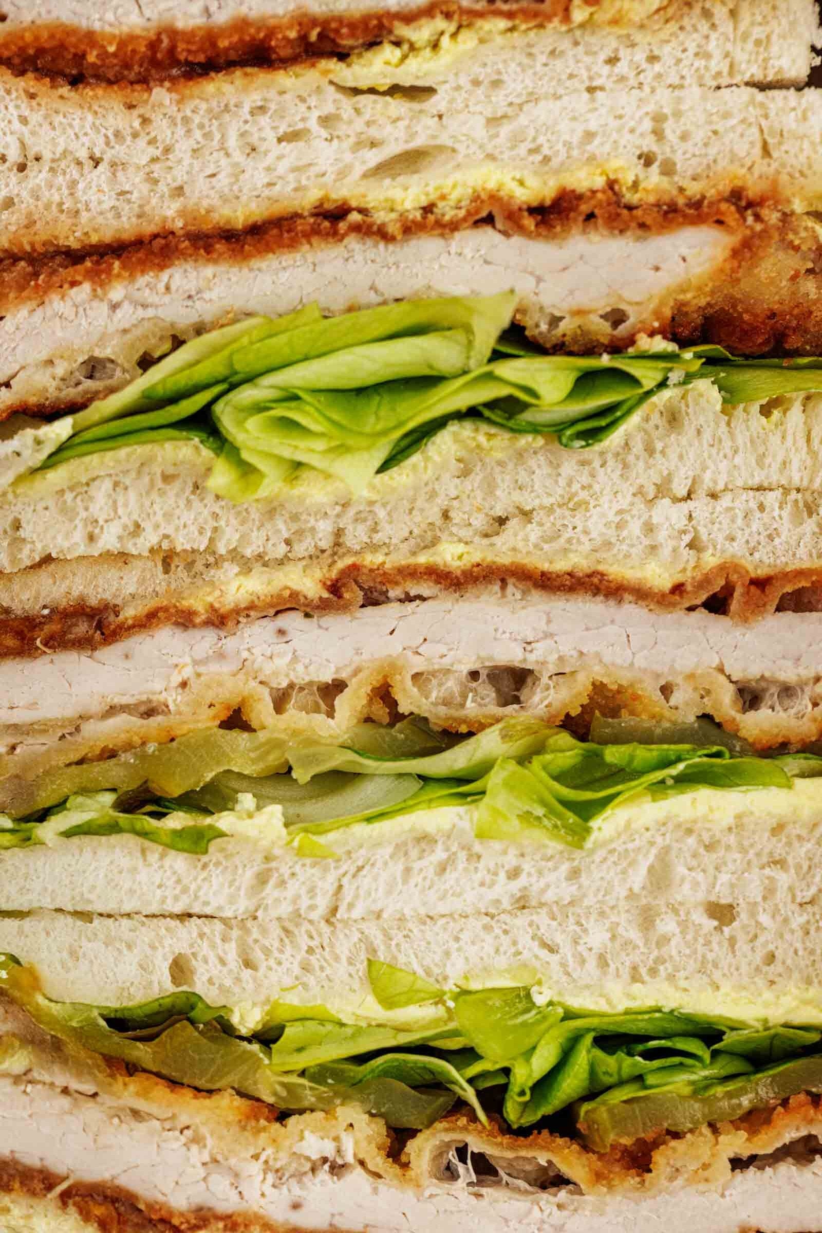 Close up cross section of stacked tonkatsu sandwiches: breaded pork cutlet, Kewpie mayo, chopped greens and pickle slices between crustless bread.